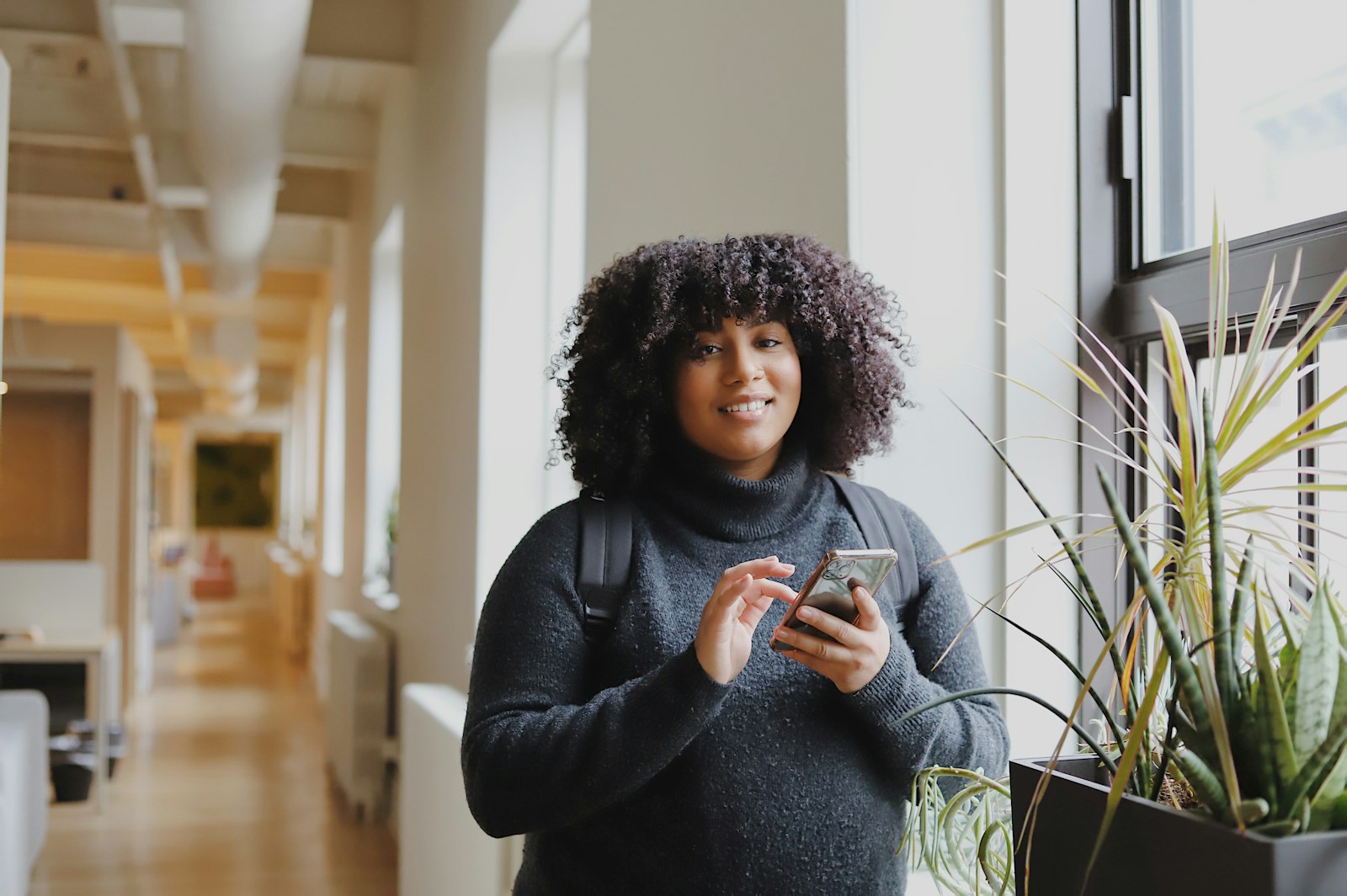 A young Black woman with natural hair smiling as she uses her smartphone by a window